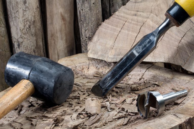 Stechbeitel mit Holzhammer und Forstnerbohrer auf einem Holzstamm – typisches Werkzeug für präzise Holzbearbeitung und Stemmarbeiten.