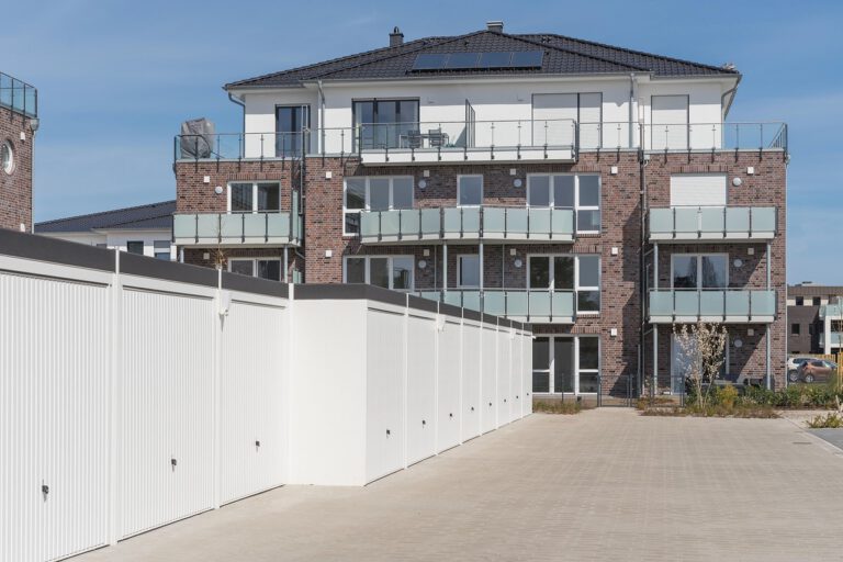 Bauabnahme Neubau: Mehrfamilienhaus mit roter Klinkerfassade, Glasbalkonen und weißen Einzelgaragen im Vordergrund