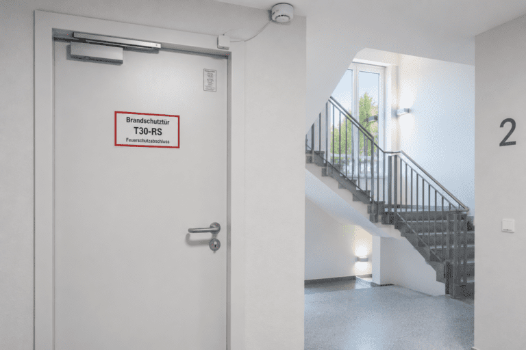 Moderne T30-RS Brandschutztür mit Kennzeichnungsschild und Rauchmelderkontakt in einem hellen Treppenraum gemäß DIN 4102 Brandschutz.